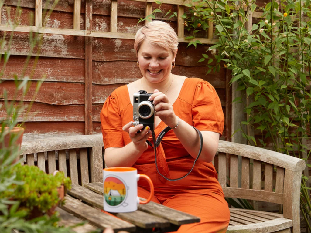 Sell on TikTok with Printify A woman with short hair, wearing an orange dress, takes a picture of a colorful mug in a garden setting.