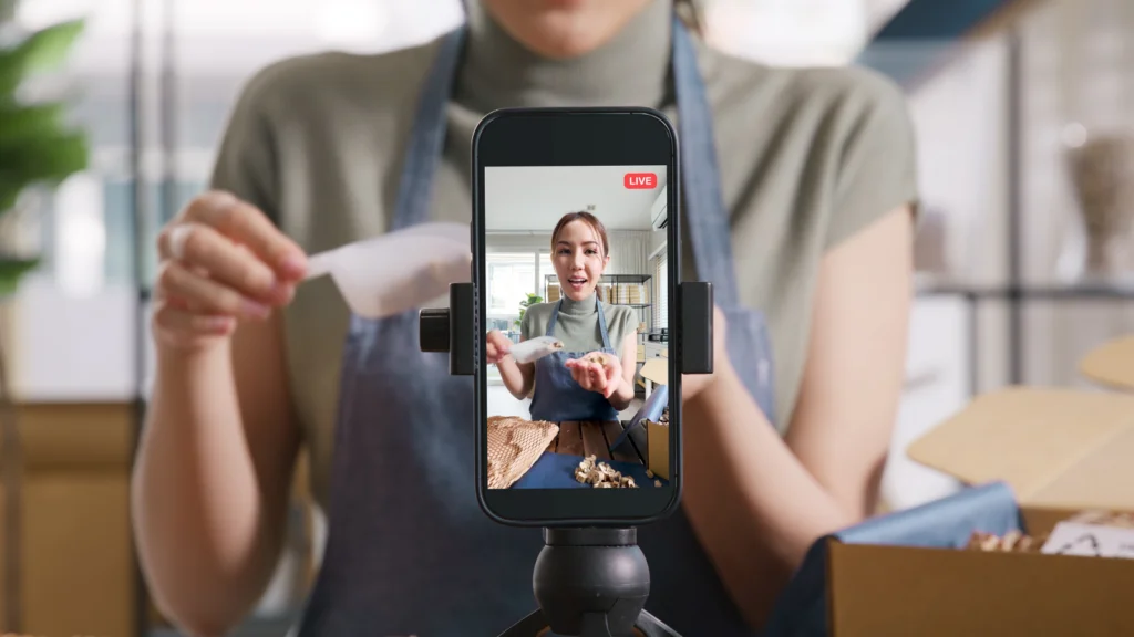 A person wearing an apron livestreams through a smartphone, enthusiastically demonstrating a craft project in a cozy, well-lit room.