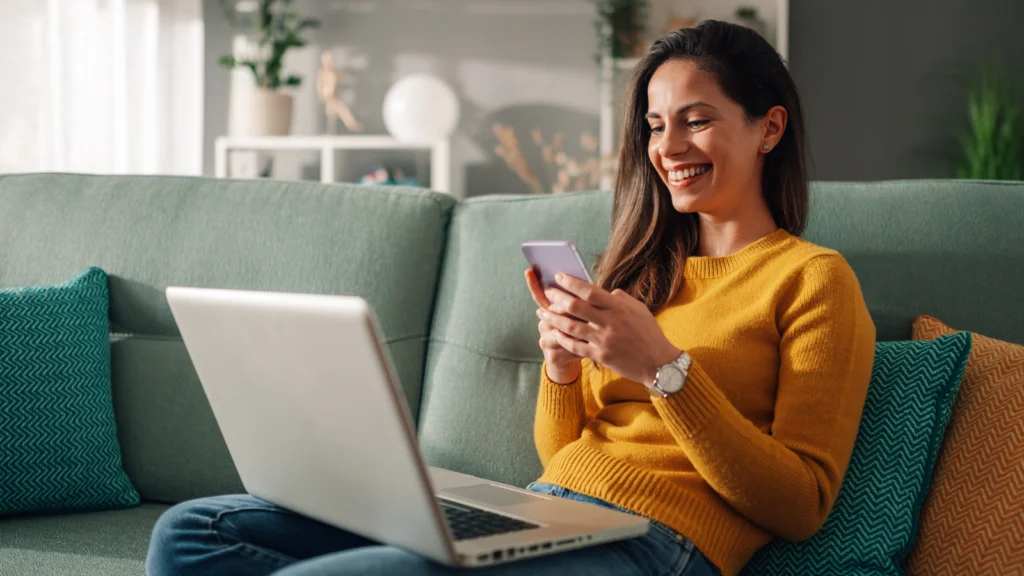 TikTok Ads metrics every eCommerce brand should track A smiling woman in a yellow sweater sits on a teal sofa, holding a smartphone in front of a laptop.