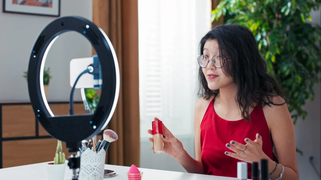 TikTok Shop affiliate program plan types A woman in glasses presents make-up products during a livestream, using a smartphone on a stand with a ring light.