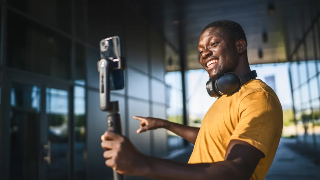 Use viral TikTok hooks to get more followers A smiling man with headphones in a yellow shirt holds a smartphone on a selfie stick, gesturing and looking at the camera.