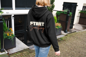 A backshot of a woman wearing a black hoodie with a “Start Something” text print in urban scenery.