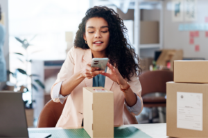 A woman with curly hair wearing a light blazer is sitting at a desk in an office, taking a photo of a package with her smartphone.