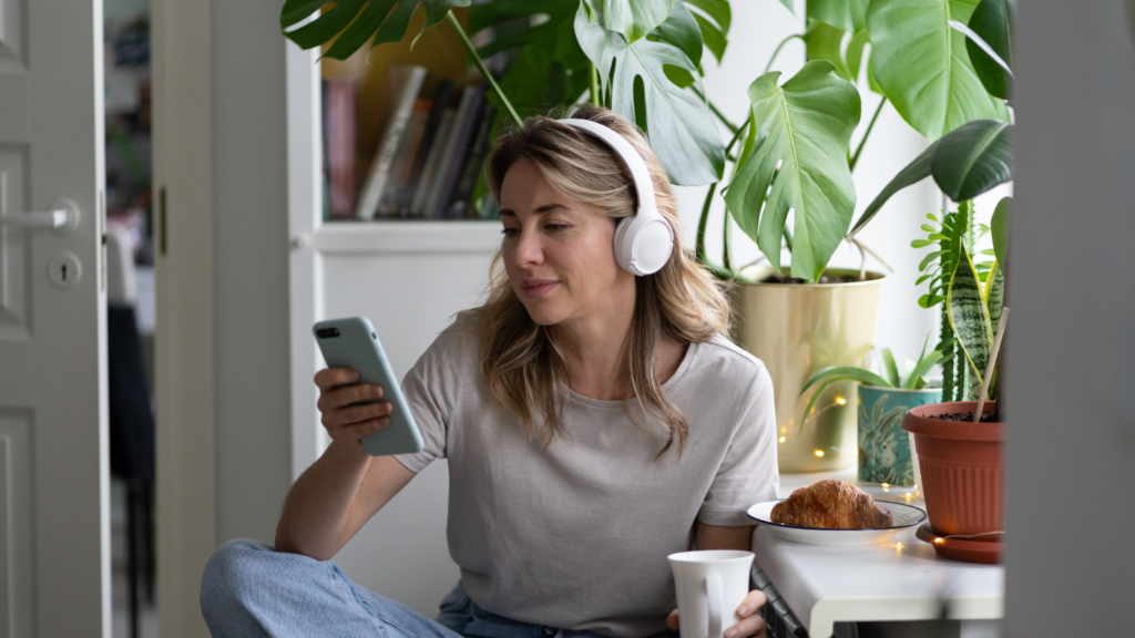 01 Individual vs business registration A woman is sitting in a kitchen, wearing headphones, drinking from a mug, and scrolling through her phone.