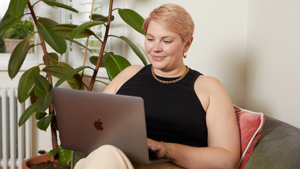 01 Why open a Printify Pop Up Store A smiling woman with short hair sits in a chair with a laptop, learning about the new Printify Pop-Up Store tools and features.
