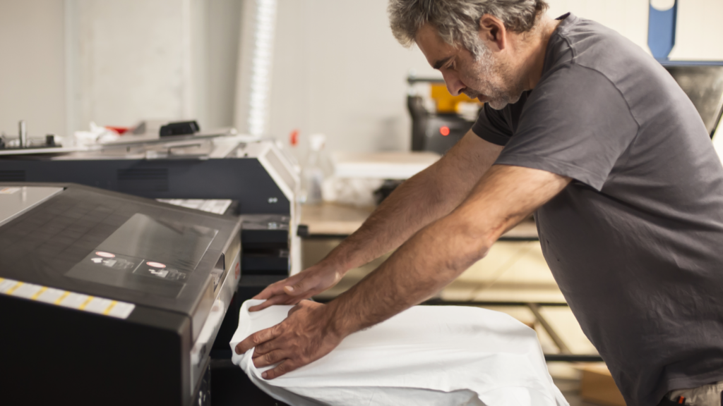A man is working on an industrial printer, preparing an organic t-shirt for printing.