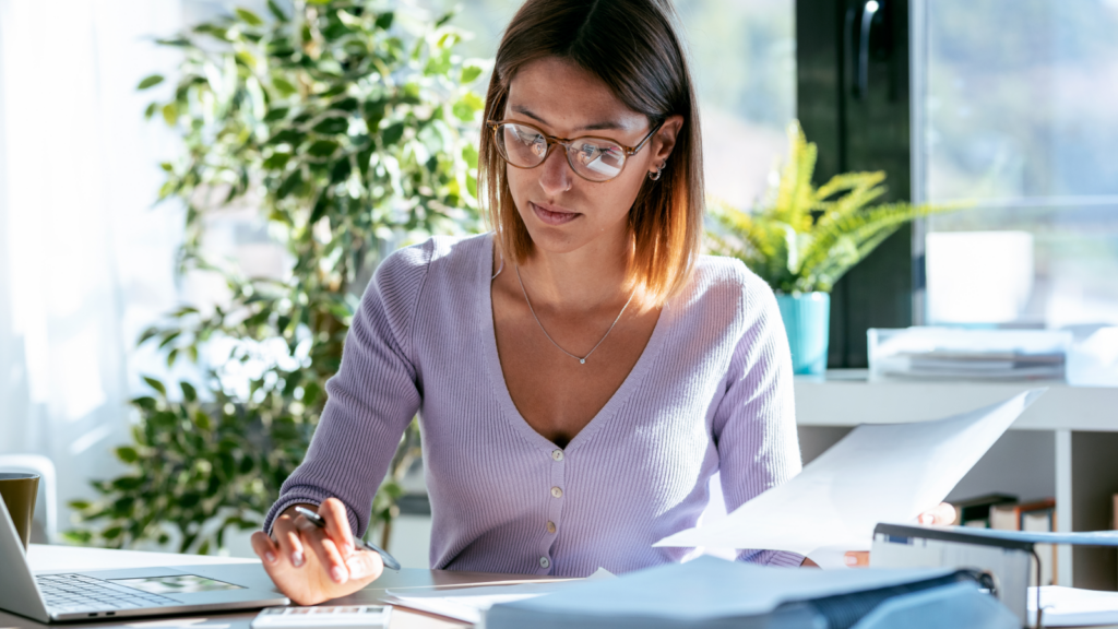 02 What this means for your costs A woman in glasses is sitting at an office desk, working with papers and using a calculator. A laptop is nearby.