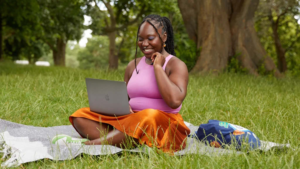 03 Explore the new tools and features yourself A smiling woman in a pink top and orange skirt is sitting on the grass in a park with a laptop.