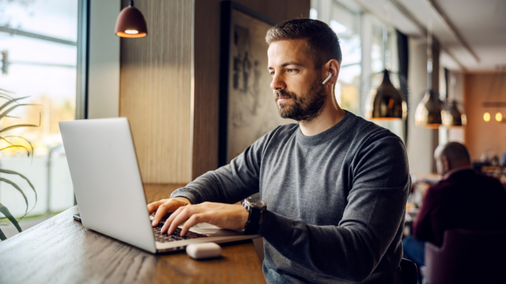 03 How to stay exempt A man with a beard, wearing a dark-blue sweater, is sitting in a cafe with a laptop.