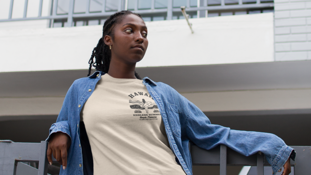 A woman is wearing a cream t-shirt with a left-chest logo, and a denim shirt. Urban scenery in the background.