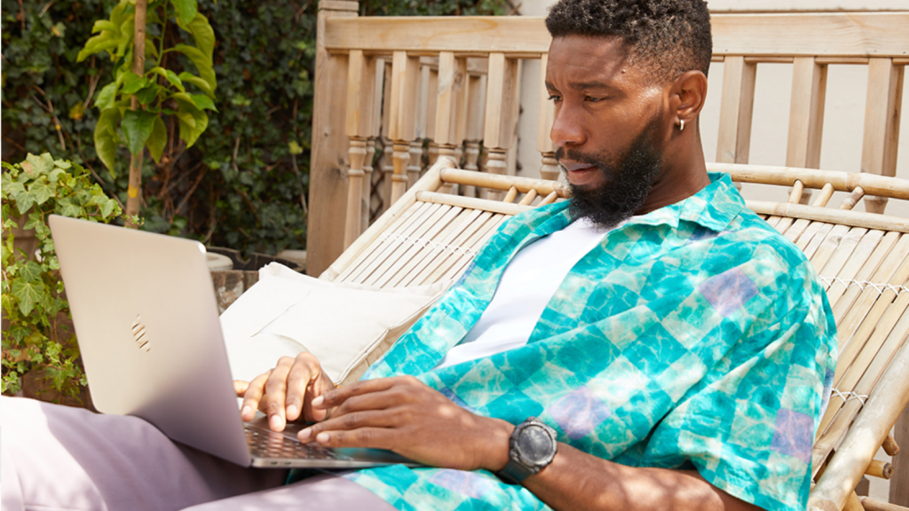 04 What Printify is doing A man in a colorful blue shirt is sitting outside with a laptop, reading the latest news on EU customs duty.