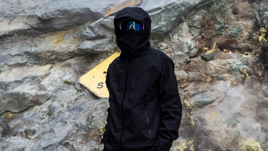 A person in visor glasses wearing a black gorpcore-themed suit. Large mountain rocks in the background.