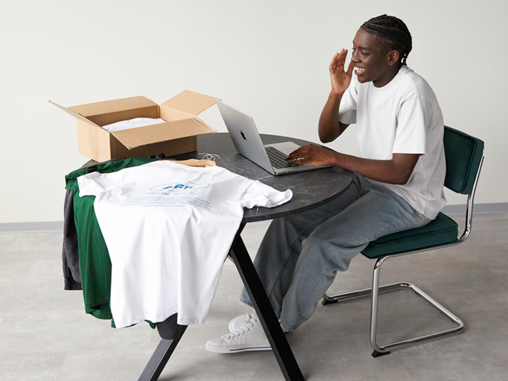 07 How TikTok Shop works with Printify A smiling young man sitting at a table with a laptop and different t-shirts nearby.