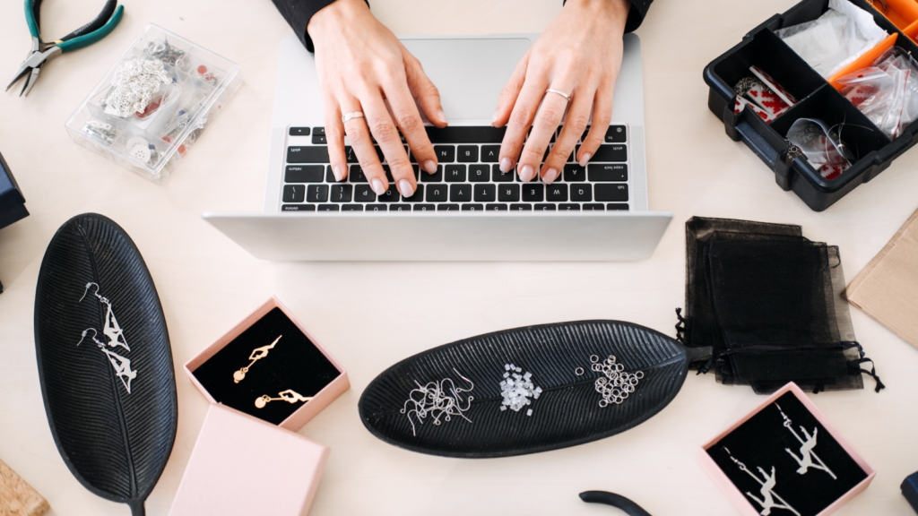 4 Etsy search terms reveal what shoppers actually type A woman sits at a table, using a laptop surrounded by an assortment of jewelry pieces.