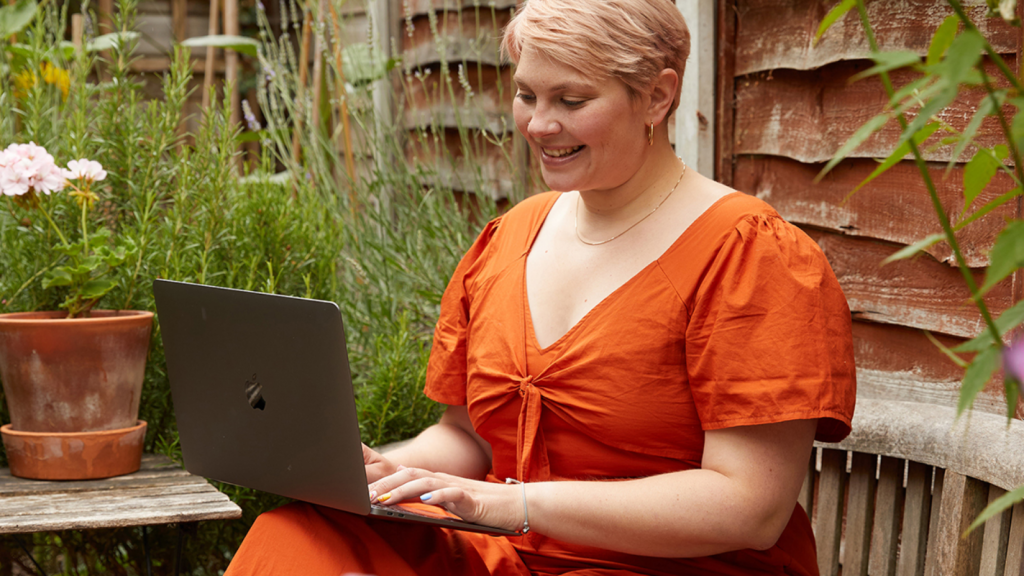 How to build a sustainable clothing brand A smiling woman in an orange dress is sitting in a home garden with a laptop, searching for eco-friendly t-shirt printing options.
