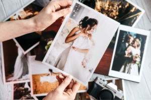 Hands holding a wedding photo of a smiling bride in a white dress, surrounded by scattered wedding pictures and a vintage camera on a table.