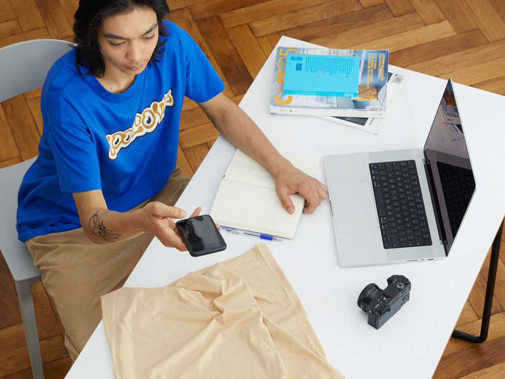 A young man taking a picture of a beige t-shirts with his smartphone. Laptop, camera and notebook are placed around the table.