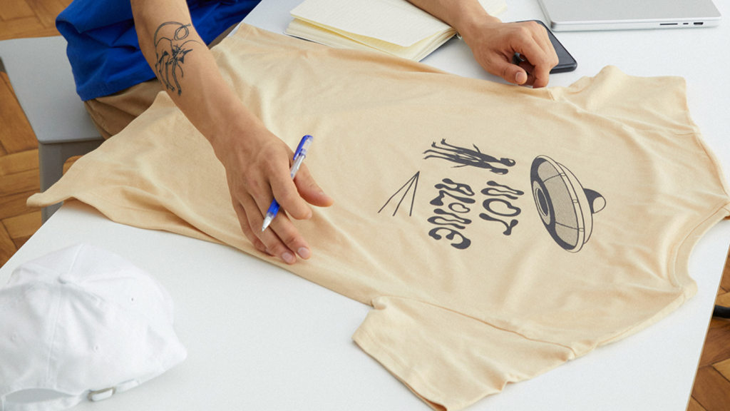 Select your t shirts A person’s hands are above a beige alien-themed “Not alone” t-shirt.
