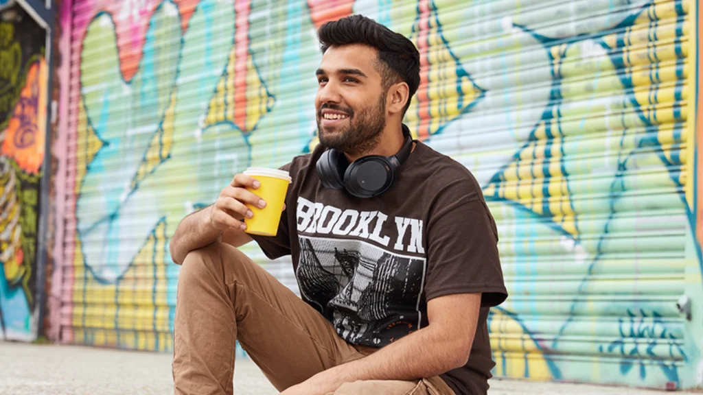 A smiling man is sitting outside with a takeaway coffee cup, wearing a brown “Brooklyn” t-shirt. Graffiti in the background.