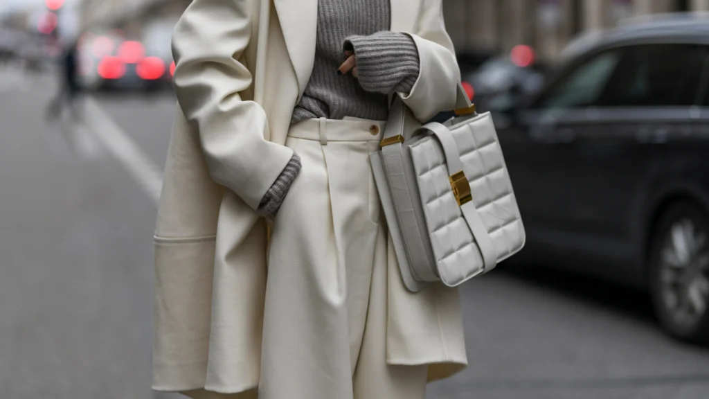 A person wearing an elegant cream coat and pants, a gray sweater, and holding a stylish quilted handbag on a city street.