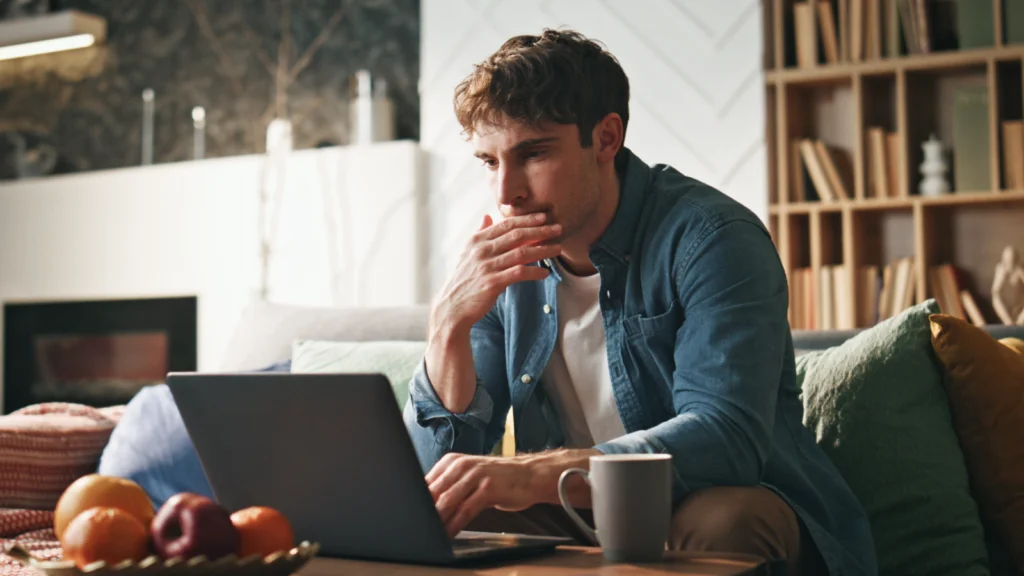 Track real ROI instead of chasing views A man in a denim shirt is sitting on a sofa, focused on a laptop in front of him, searching for advanced TikTok affiliate marketing strategies.