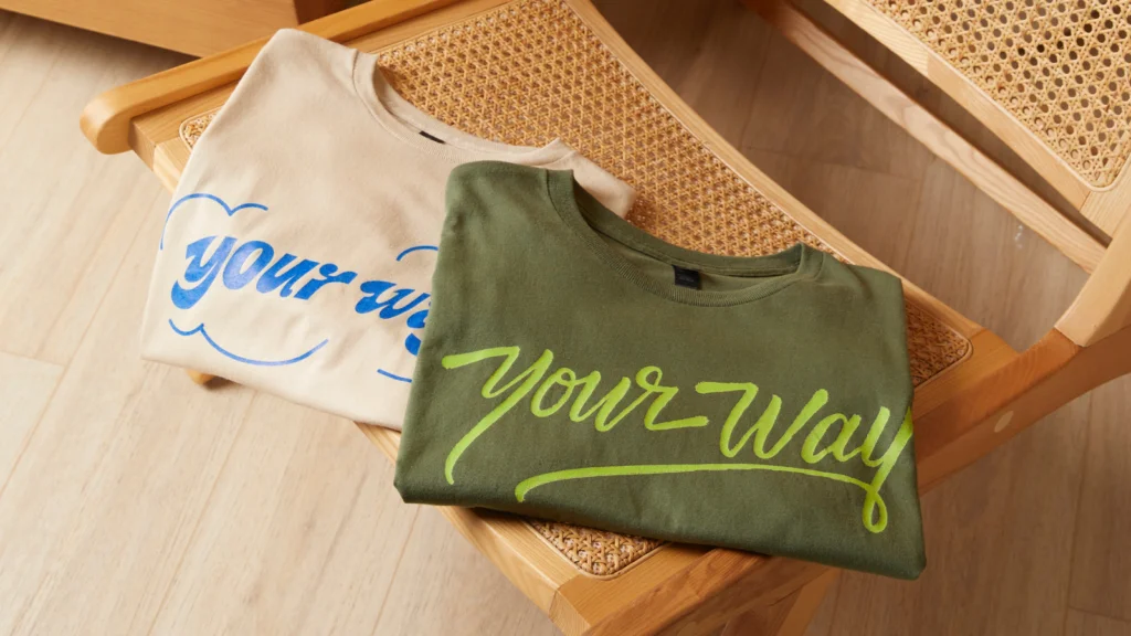 Upload your final designs Two folded “Your way” t-shirts on a wooden chair. One t-shirt is cream with blue text, the second t-shirt is khaki with green text.