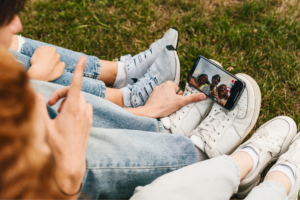 Three people sit on grass, legs close together, with a smartphone on one of the people’s shoes, taking a selfie.