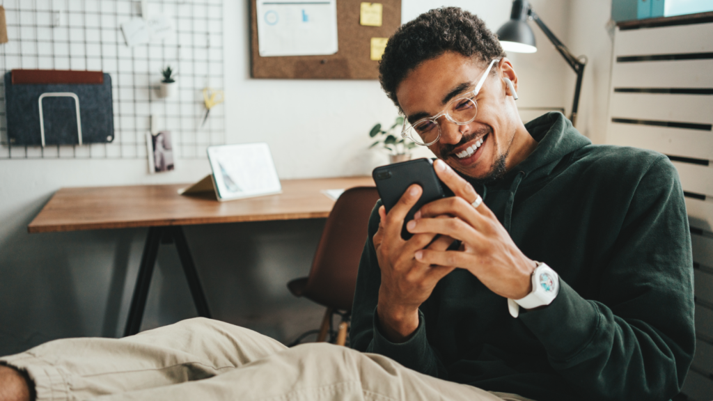 01 TikTok Shop ad formats A smiling man with glasses, wearing a green sweater and beige pants, is sitting in an office, legs on the table, looking at his phone.