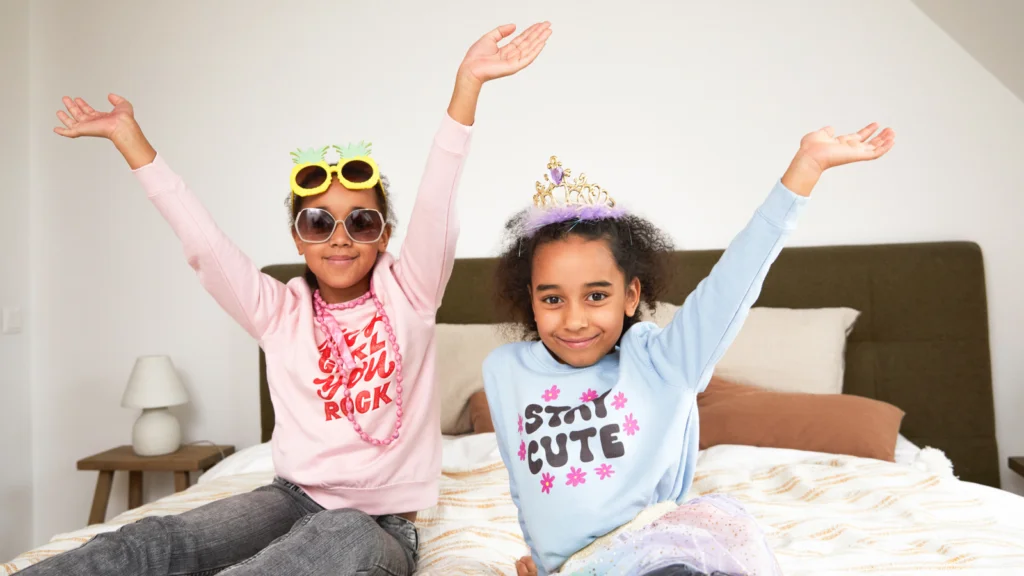 05 Childrens clothing Two young girls are sitting on a bed, wearing colorful outfits and accessories, with their arms raised in a joyful pose.