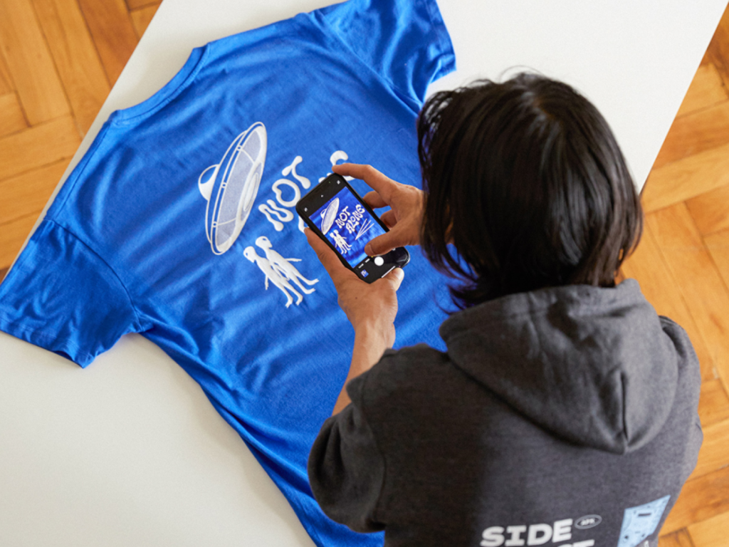 05 Sell on TikTok Shop with Printify A man is taking a picture of a blue “Not alone” alien-themed t-shirt, using a smartphone.
