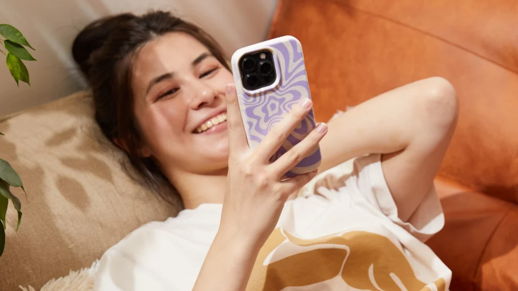 06 Phone cases A young woman lies on a couch, smiling as she looks at her phone. She has long hair and is wearing a casual t-shirt.