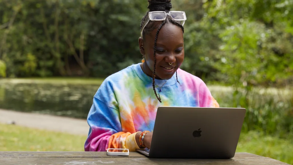 07 How to find best selling items on Etsy A young woman sitting at a table outdoors, wearing a colorful tie-dye sweatshirt, using a laptop, and smiling.
