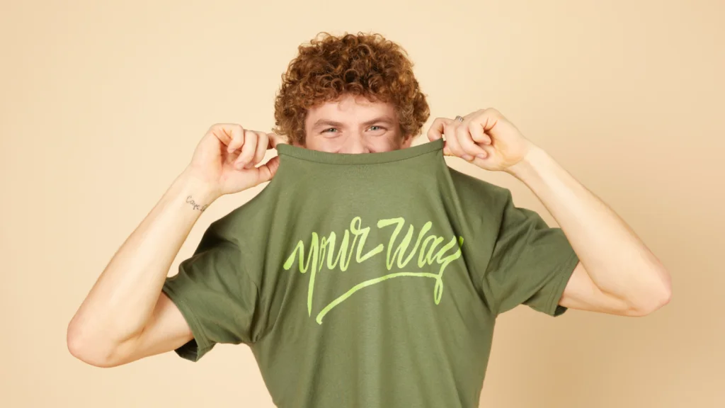 Apparel thumbnails A young man with curly hair, wearing a green “Your way” t-shirt, pulls it halfway over his head. Light yellow background.