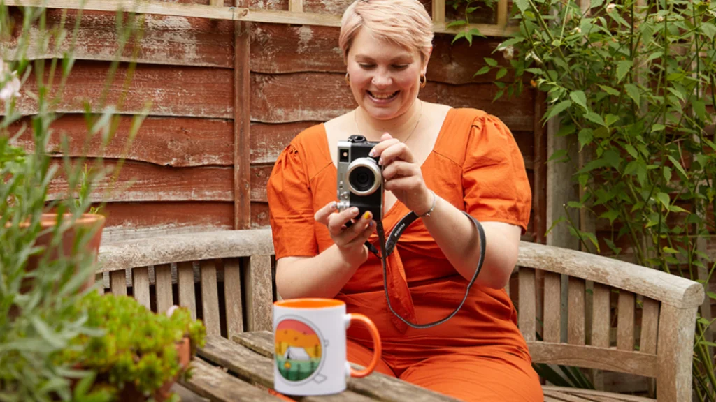Promote your mug printing business A woman with short blonde hair, wearing an orange dress, is taking a picture of a colorful mug using a digital camera. Home garden in the background.