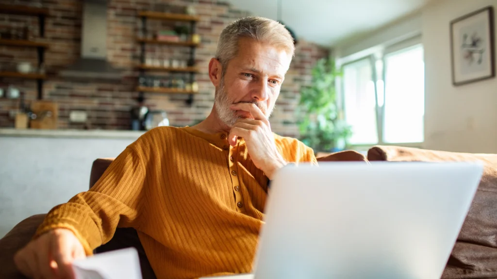 Why create personalized mugs A man sitting on a couch, looking thoughtfully at a laptop in a bright and cozy room.